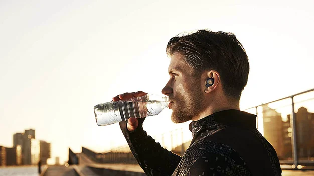 Runner wearing running headphones while drinking from a bottle of water in front of a city skyline at dawn